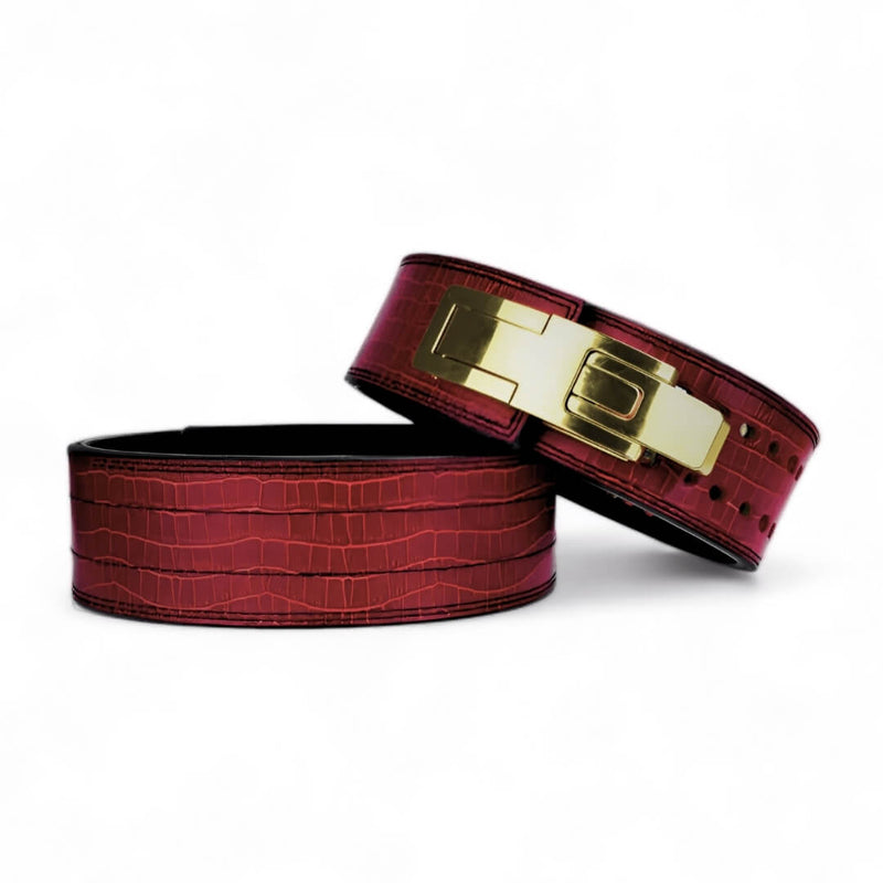 Two burgundy croc-grain Kobra lever belts on a white background, one showing its gold lever buckle.