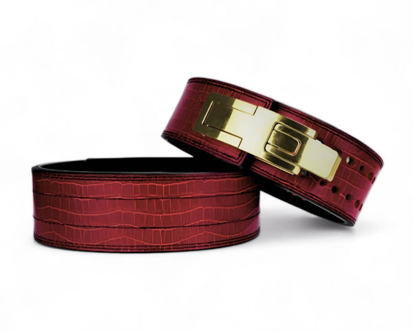 Two burgundy croc-grain Kobra lever belts on a white background, one showing its gold lever buckle.